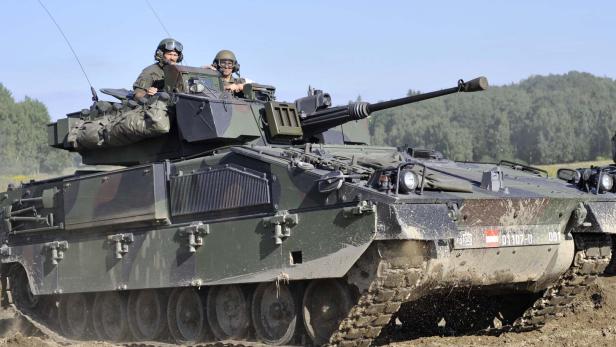 Zwei Soldaten fahren in einem gepanzerten Kettenfahrzeug mit Tarnanstrich und Kanone über einen schlammigen Untergrund.